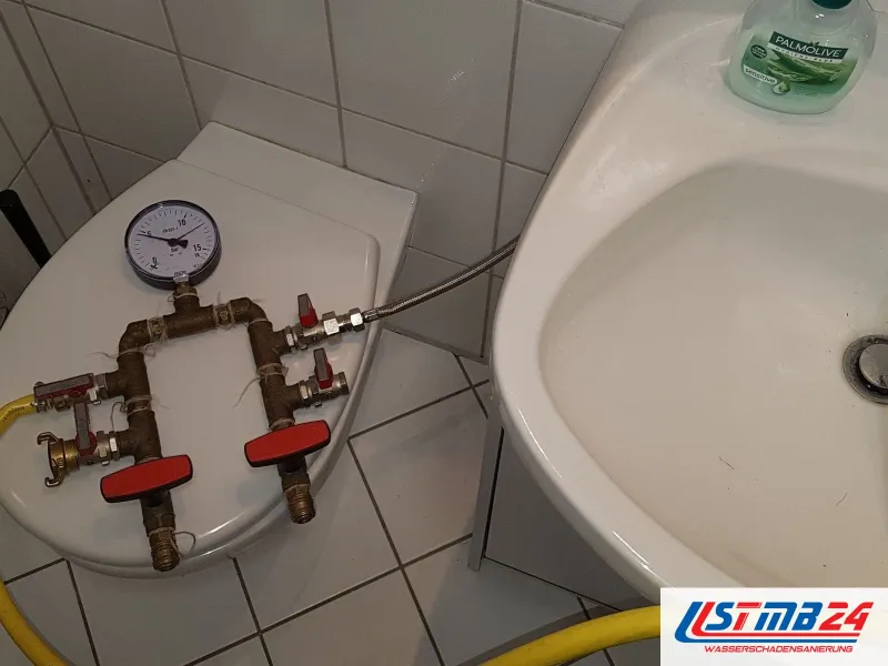 Prüfarmatur mit Manometer und roten Hähnen auf einem WC-Deckel, gelbe Schläuche führen zum Zulauf