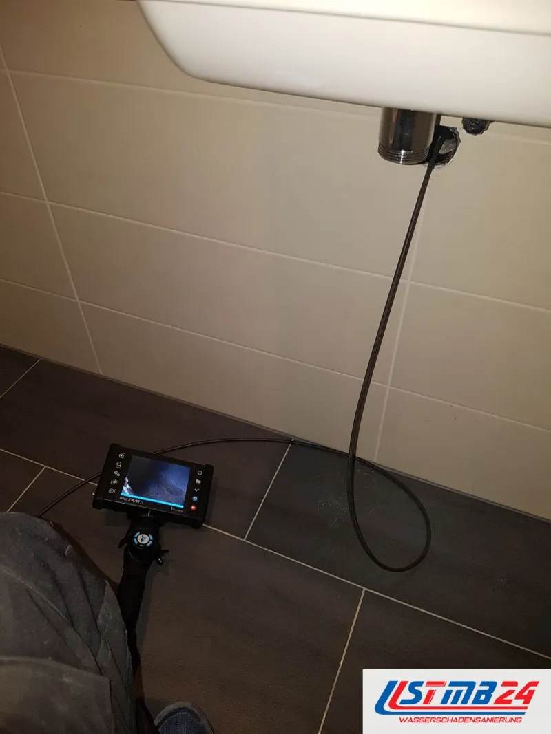 Endoskop mit eingeschaltetem Monitor auf dunklen Bodenfliesen, das Kabel führt hoch zum Anschluss unter dem Waschbecken
