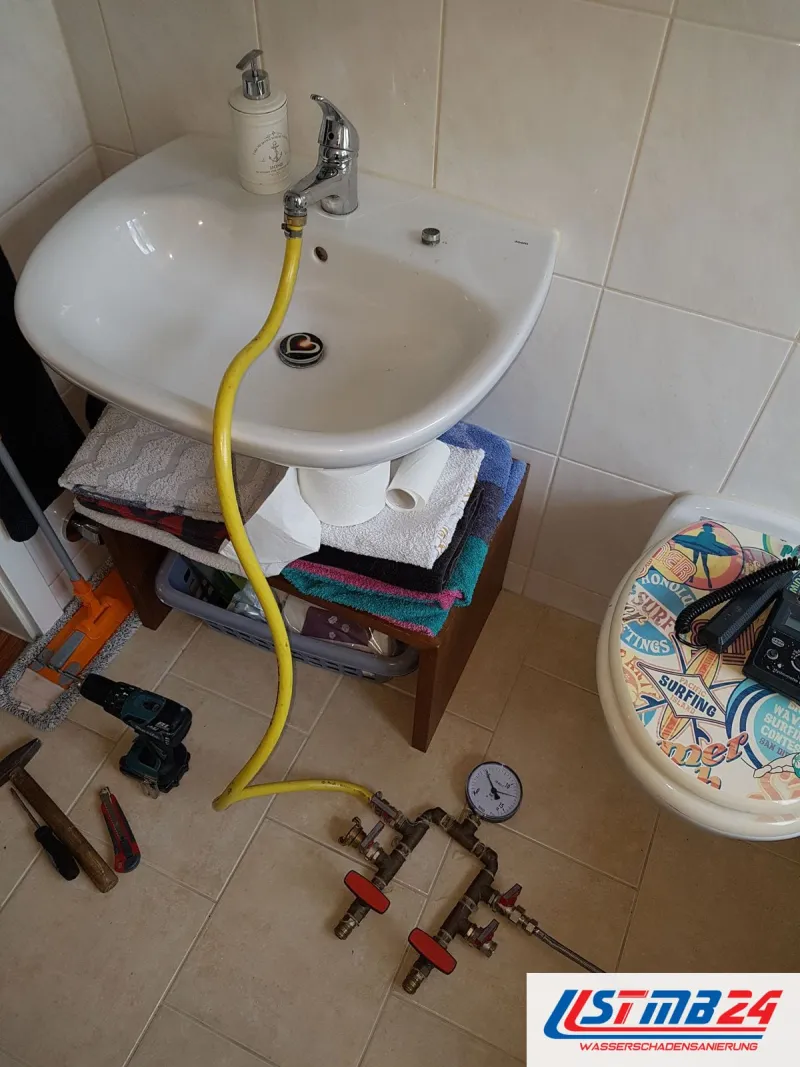 Waschbecken mit gelbem Schlauch zum Prüfgerät auf den Bodenfliesen, daneben Werkzeug und ein WC mit gemustertem Deckel