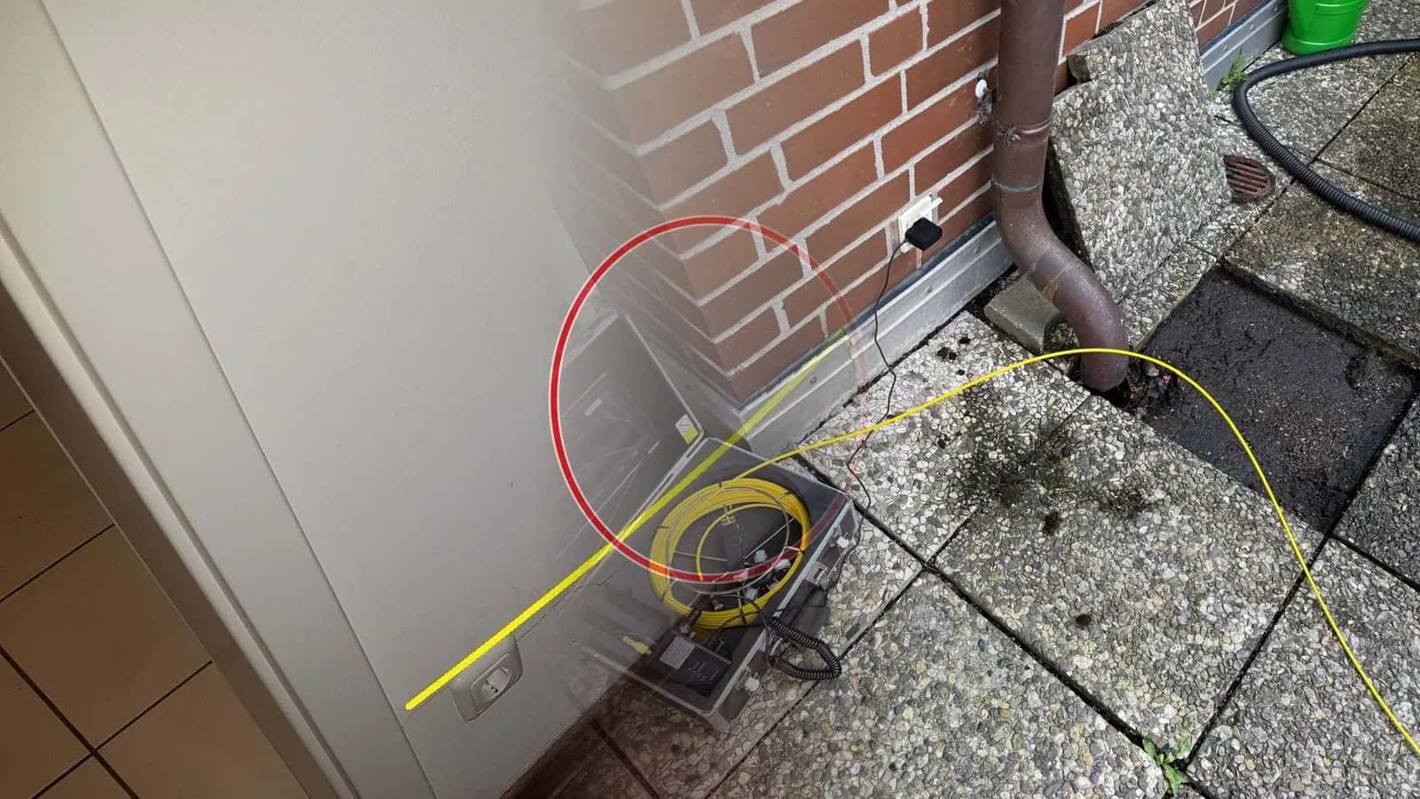 Rohrkamera im Koffer mit Monitor auf einer Terrasse, das gelbe Kabel führt in das Regenfallrohr am Klinkerhaus