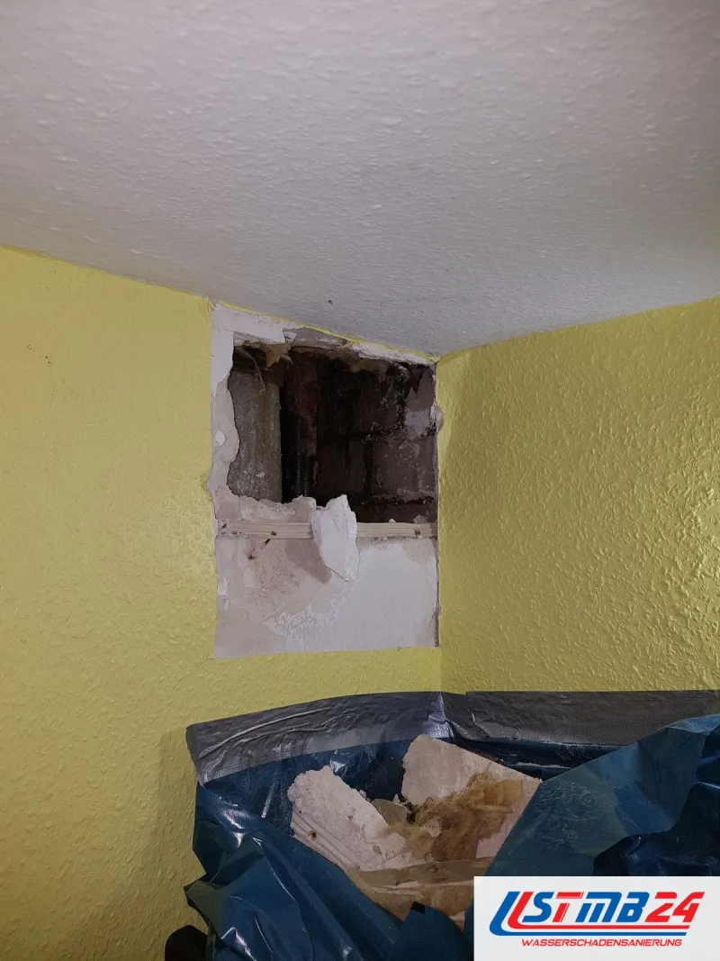 Geöffnete Wand direkt unter der Decke in einem gelb gestrichenen Raum, darunter Schutt in einem blauen Sack