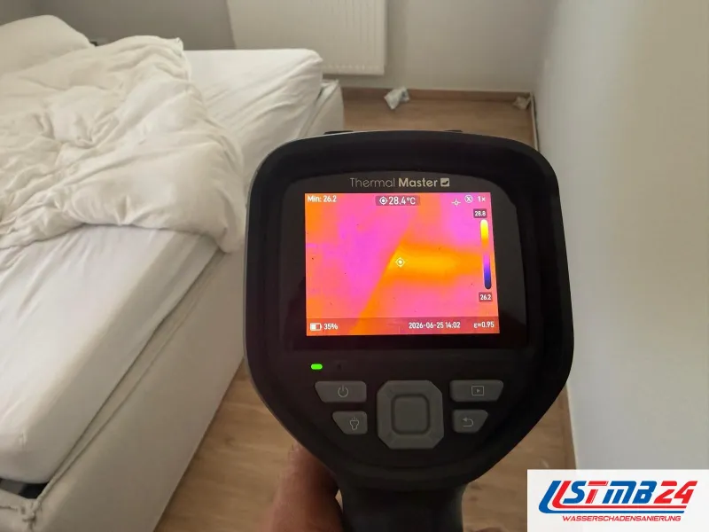 Wärmebildkamera vor einer Schlafzimmerwand, auf dem Display ein warmer gelber Streifen bei 28,4 Grad neben kühlen violetten Flächen