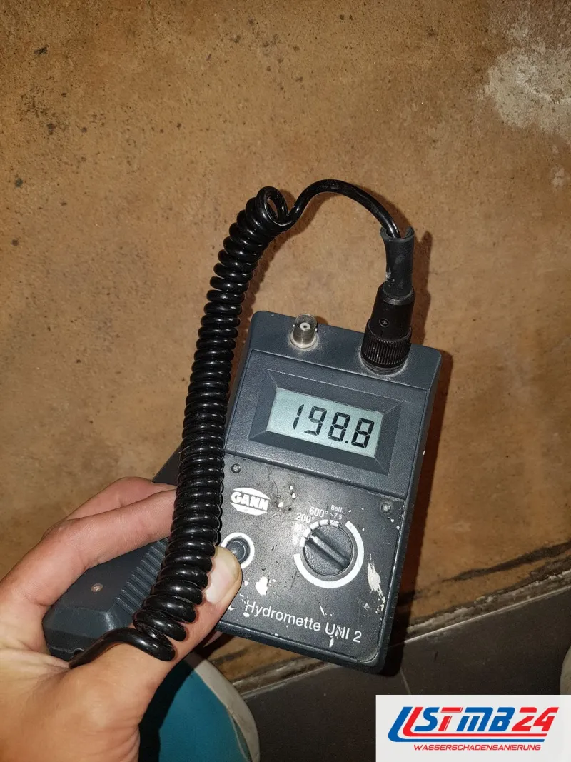 Feuchtemessgerät mit 198,8 auf dem Display vor einer nassen Wand mit dunklem Rand über dem Boden
