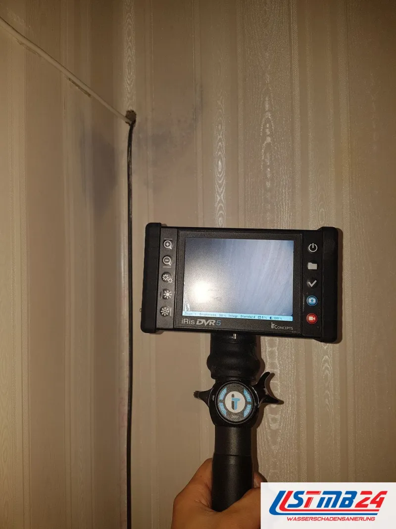 Endoskop mit Monitor vor einer gestreiften Tapete, das Kabel führt in eine kleine Bohrung neben einem feuchten Fleck