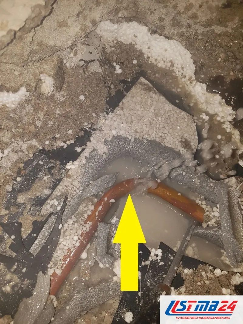 Geöffneter Estrich mit nassen Styroporkügelchen, ein Kupferrohr liegt frei, der gelbe Pfeil zeigt auf die undichte Stelle