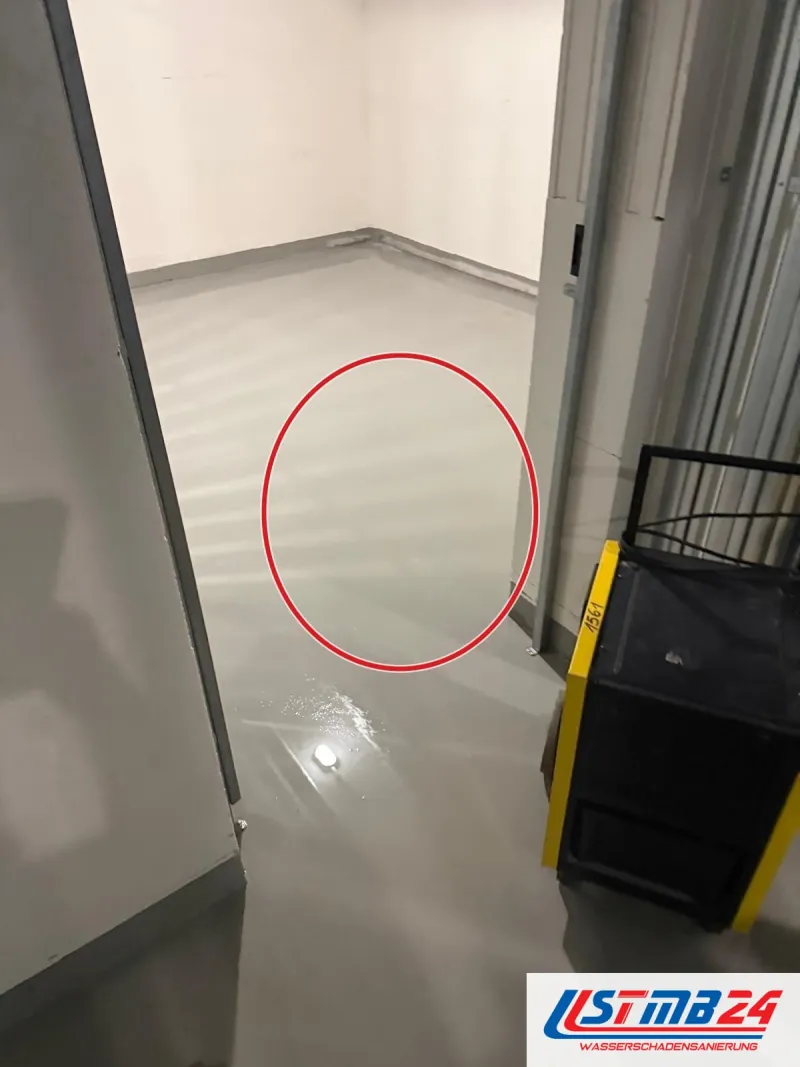 Kellerraum im Neubau, Wasser glänzt auf dem grauen Boden, im Gang ein schwarz-gelber Bautrockner, ein roter Kreis markiert die Stelle