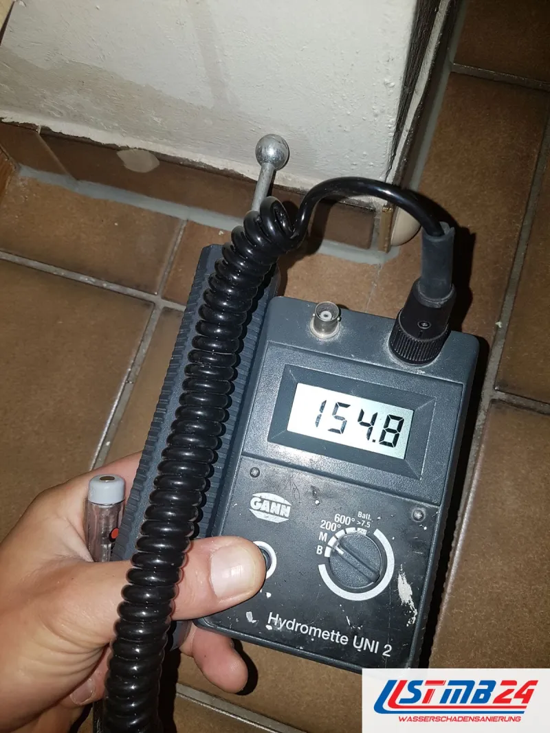 Feuchtemessgerät mit 154,8 auf dem Display vor einem gefliesten Sockel, die Sonde mit der Messkugel liegt an der Wand