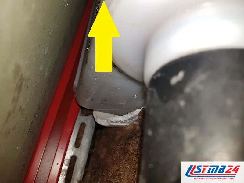 Abflussbogen hinter einer Verkleidung, ein gelber Pfeil zeigt auf die nasse Stelle