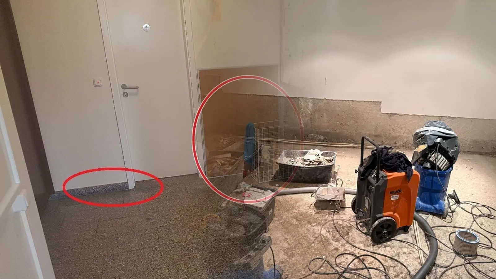 Souterrain mit orangefarbenem Bautrockner, Wassersauger und Werkzeugkisten, der Sockelputz bis Kniehöhe abgeschlagen