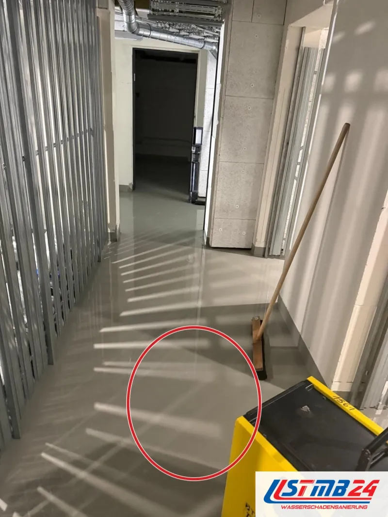 Flur mit unbeplankten Metallständern, Wasser glänzt auf dem Estrich, vorn ein gelbes Trocknungsgerät, ein roter Kreis markiert die Stelle