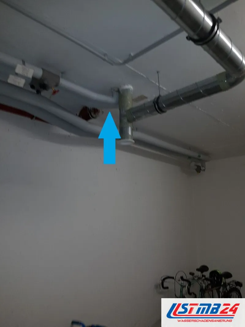 Kellerraum mit Fahrrädern, unter der Decke Wasser- und Lüftungsrohre, ein blauer Pfeil zeigt auf ein Abzweigstück