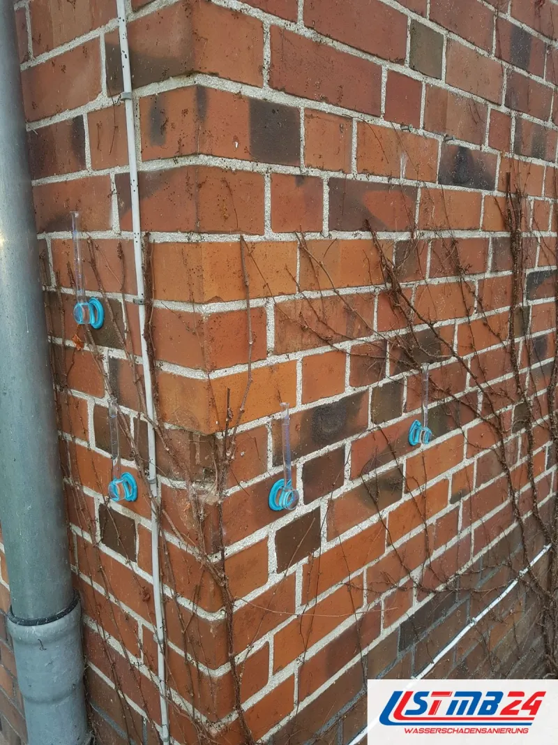Hausecke aus rotem Klinker neben einem Fallrohr, blaue Markierungen stecken an mehreren Stellen in den Fugen