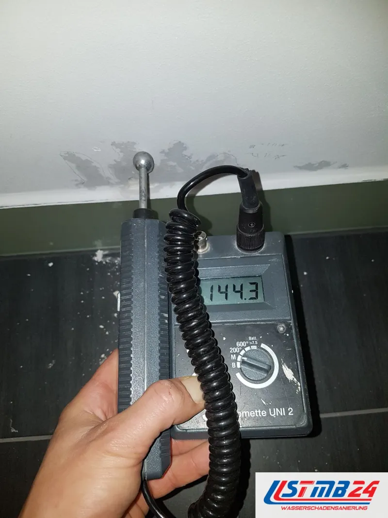 Messgerät mit Anzeige 144,3 in der Hand, dahinter eine Wand mit Flecken über dunklem Boden
