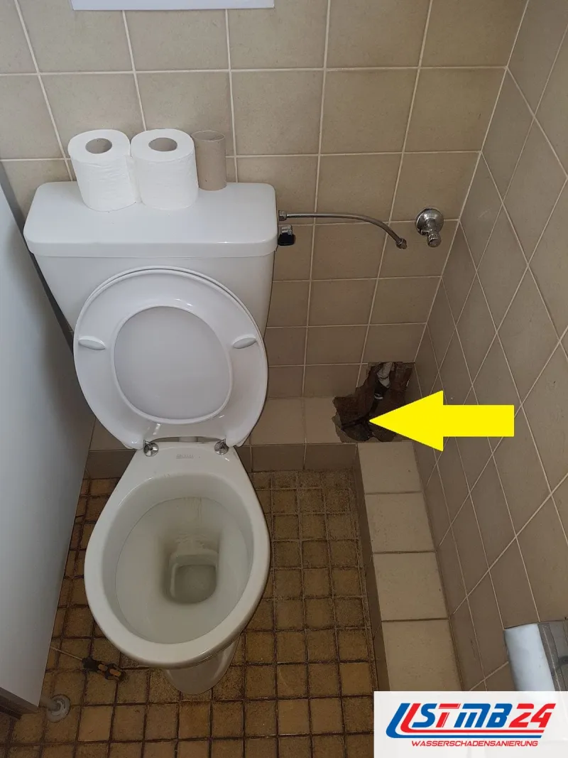 Stand-WC vor gefliester Wand, ein gelber Pfeil zeigt auf eine geöffnete Stelle neben dem Becken