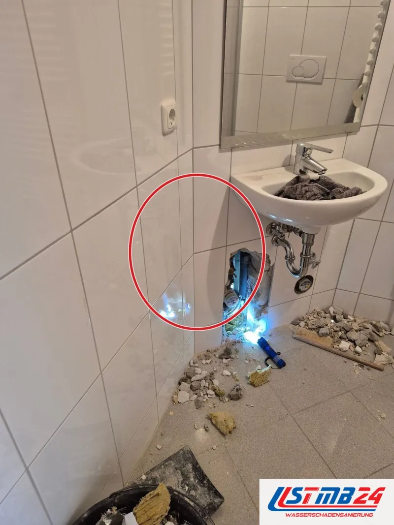 Gäste-WC mit weißen Fliesen, unter dem Waschbecken ist die Wand geöffnet, eine Lampe leuchtet hinein, ein roter Kreis markiert die Stelle