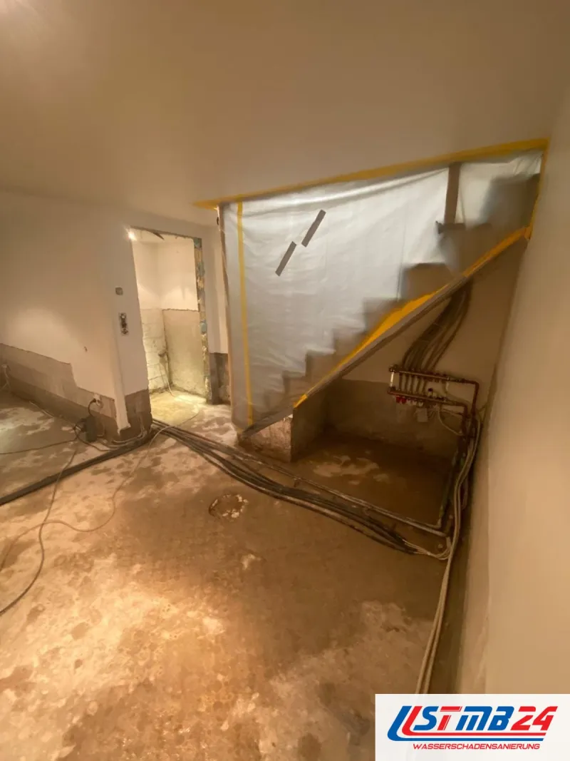 Entkernter Keller mit Staubschutzwand aus Folie vor der Treppe, Heizkreisverteiler an der Wand, Kabel und Schläuche auf dem Estrich