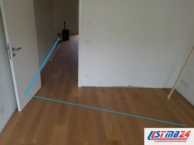 Wohnraum mit blauer Markierungslinie auf dem Parkett von der Tür zur Wand, im Flur dahinter ein laufendes Trocknungsgerät