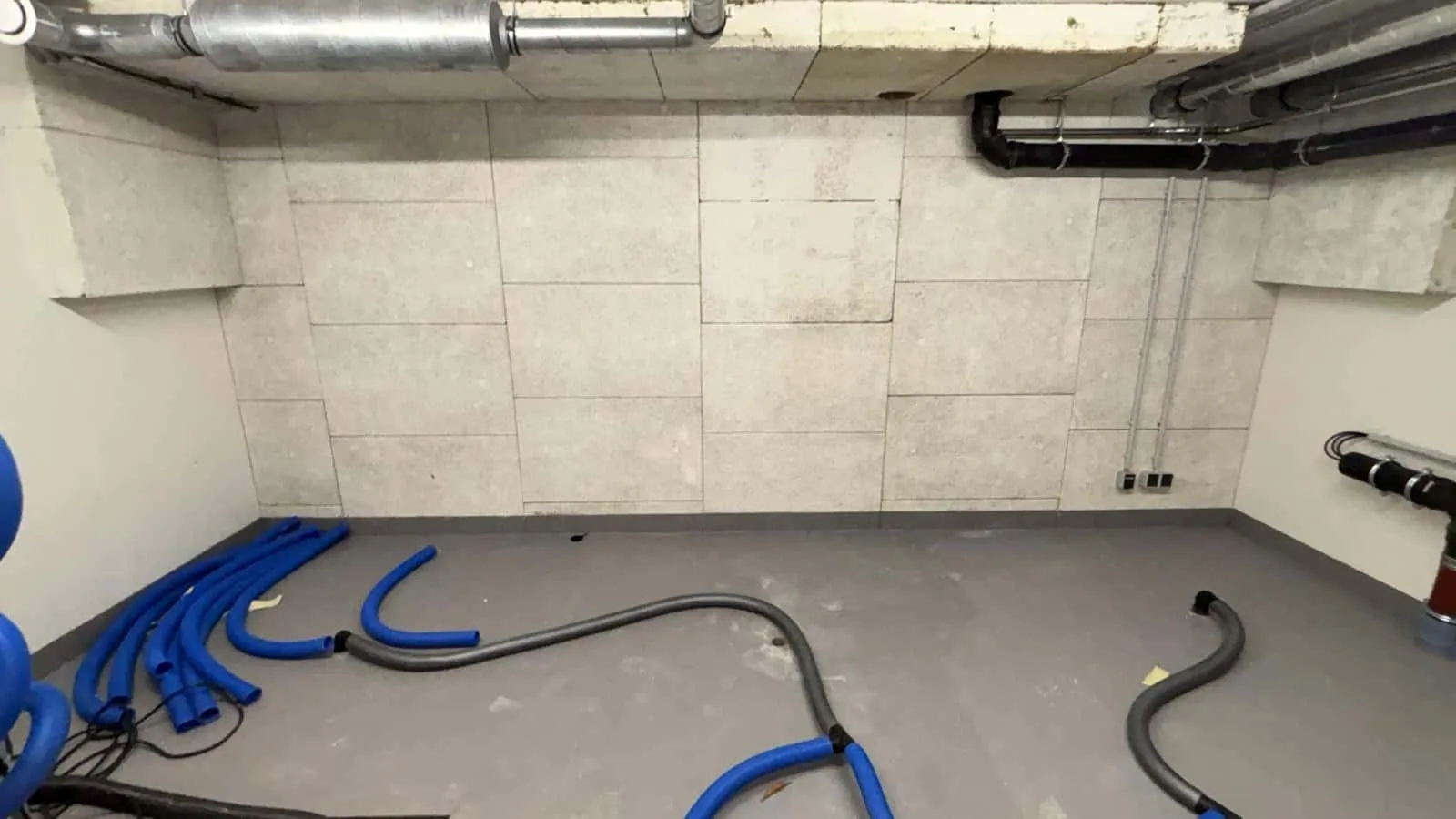 Kellerraum nach einem Wasserschaden: blaue Trocknungsschläuche führen in Bohrungen an der Wand