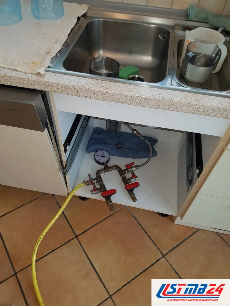 Prüfarmatur mit Manometer und zwei roten Kugelhähnen unter einer Spüle, gelber Schlauch zum Anschluss, Terrakottafliesen