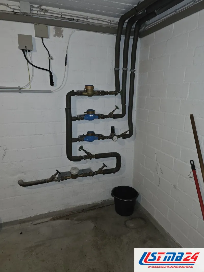 Hausanschluss mit drei Wasserzählern übereinander, gedämmte Leitungen und Absperrventile an weißer Kellerwand, Eimer darunter