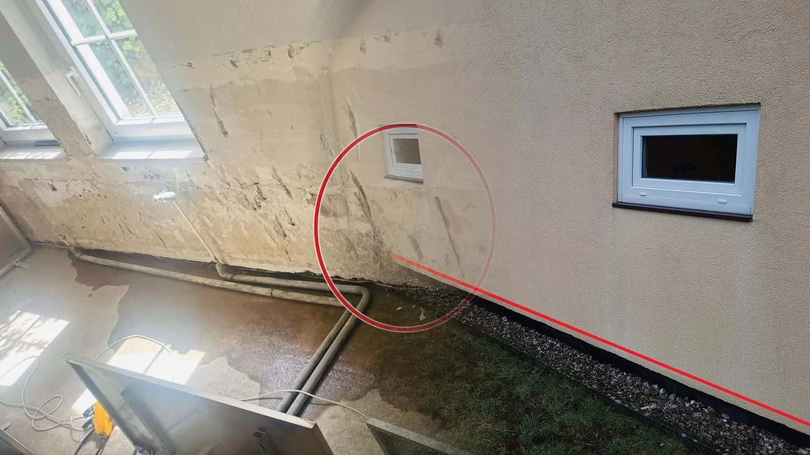 Hauswand mit zwei Kellerfenstern über einem Kiesstreifen, eine rote Linie am Sockel markiert die feuchte Zone