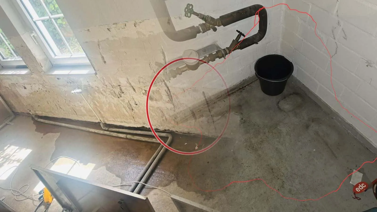 Wasserzähler und Absperrventile in einer Kellerecke, davor die rot umrandete nasse Stelle am Boden und ein Eimer