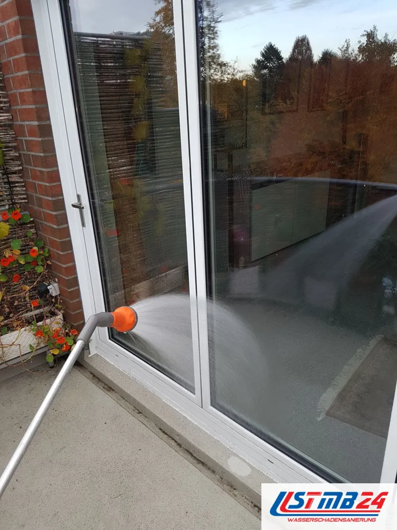 Fenster und Türen: Beregnungstest an der Balkontür, eine orangefarbene Brause am Stab sprüht Wasser auf Glas und Rahmen
