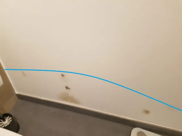 Dokumentation: blaue Linie im Foto markiert die Feuchtegrenze über braunen Wasserflecken an der Wand eines WCs