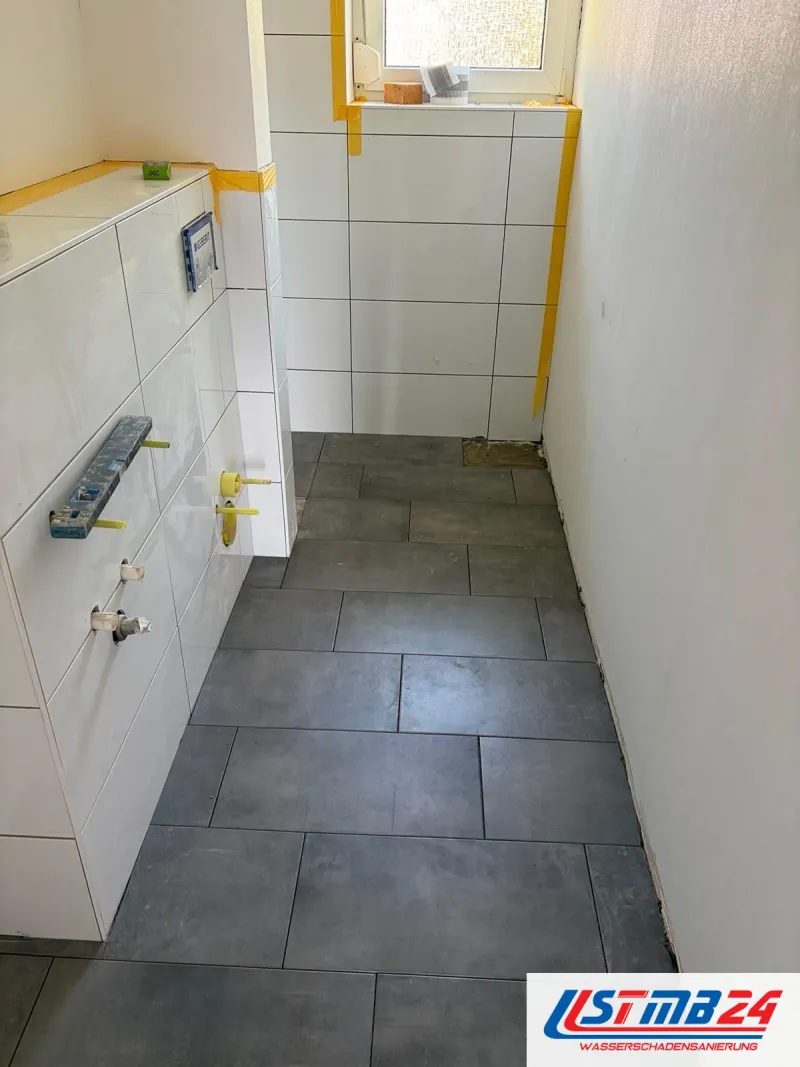 Bad im Wiederaufbau: Vorwand und Wände weiß gefliest, dunkle Bodenfliesen im Großformat, Wasseranschlüsse mit gelben Stopfen verschlossen
