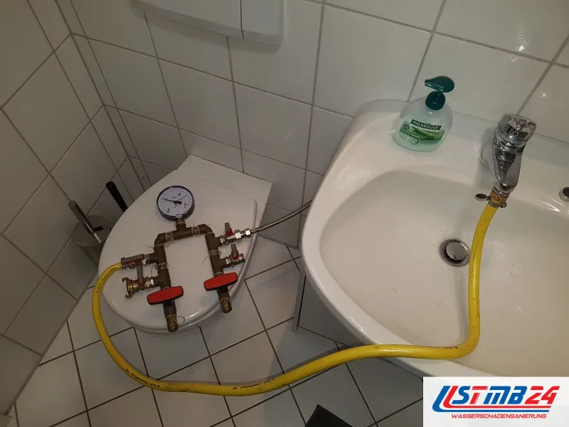 Leckageortung mit Druckprüfung: Prüfarmatur mit Manometer und zwei roten Kugelhähnen auf dem WC-Deckel, Zulauf über gelben Schlauch