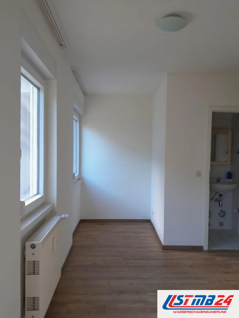 Maler- und Lackierarbeiten fertig: Zimmer mit zwei Fenstern, weiße Wände, Heizkörper, Boden in Holzoptik, Blick ins Bad