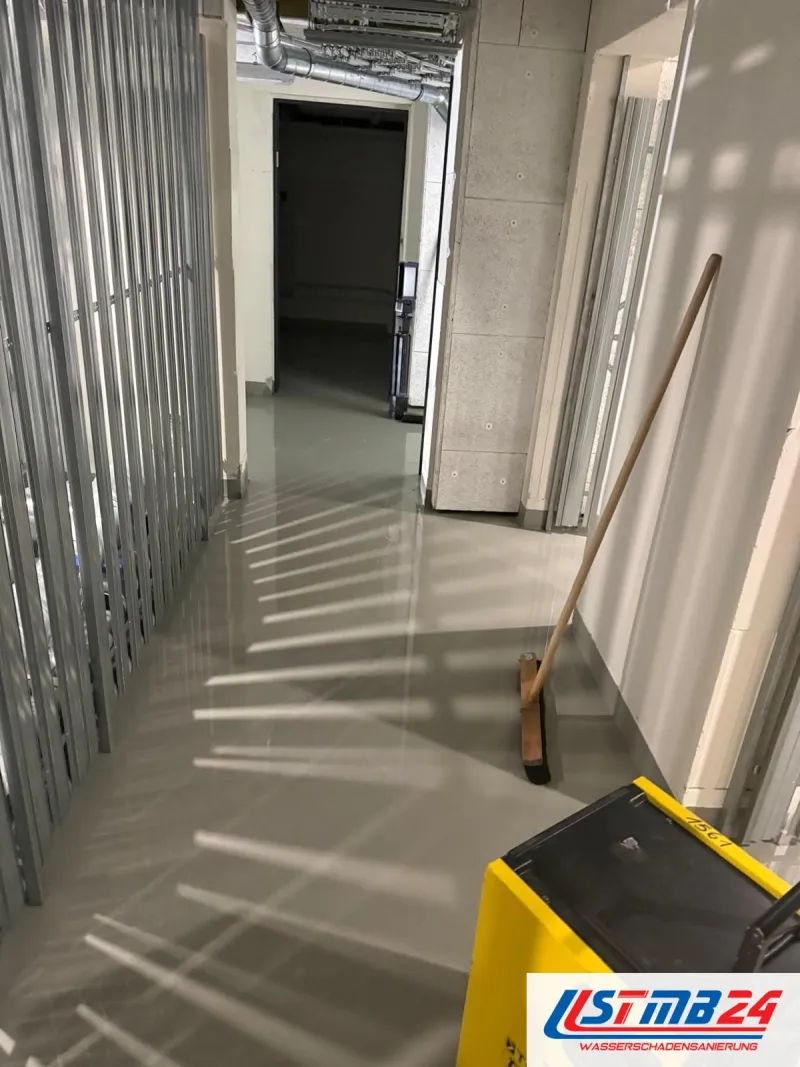 Neubautrocknung im Rohbau: Flur mit unbeplankten Metallständern, Wasser glänzt auf dem Estrich, vorn ein gelbes Trocknungsgerät