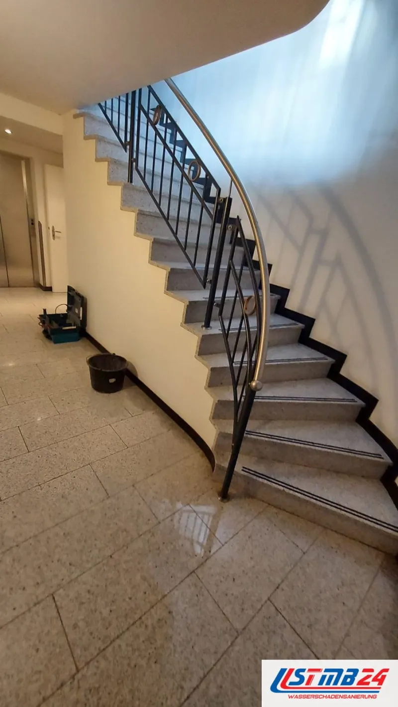 Wasserschaden in Geesthacht: Treppenhaus mit geschwungener Treppe, am Fuß Eimer und Werkzeugkoffer, der Steinboden glänzt nass