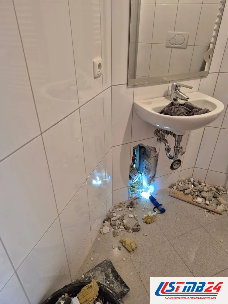 Badsanierung in Geesthacht: Gäste-WC mit weißen Fliesen, unter dem Waschbecken ist die Wand geöffnet, eine Lampe leuchtet hinein, Schutt am Boden