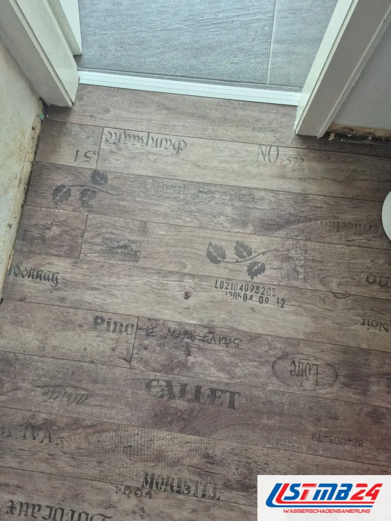Bodenleger in Geesthacht: Vinyl in Holzoptik mit Weinkisten-Aufdruck vor der Badtür, am Wandfuß Feuchteränder