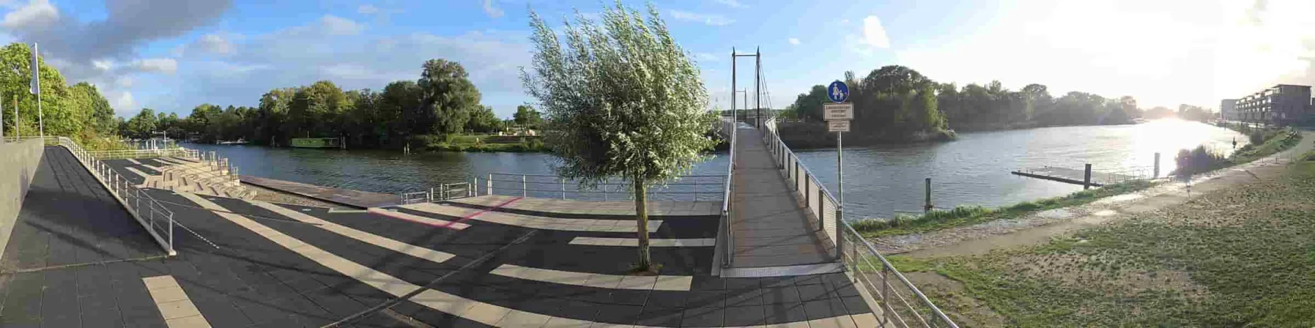 Küchenmontage in Geesthacht: Uferpromenade mit jungem Baum und Fußgängerbrücke an einem sonnigen Tag