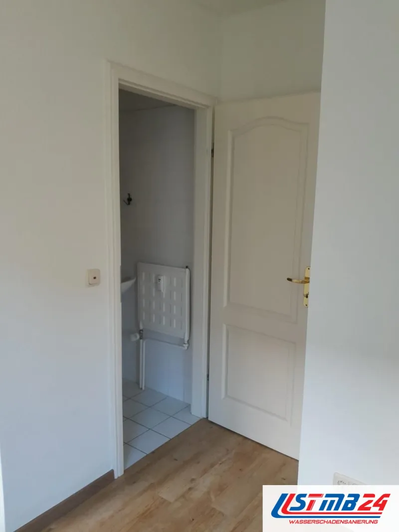Nach der Sanierung in Grömitz: weiß gestrichenes Zimmer mit Holzboden, durch die Tür Blick ins Bad mit Duschklappsitz