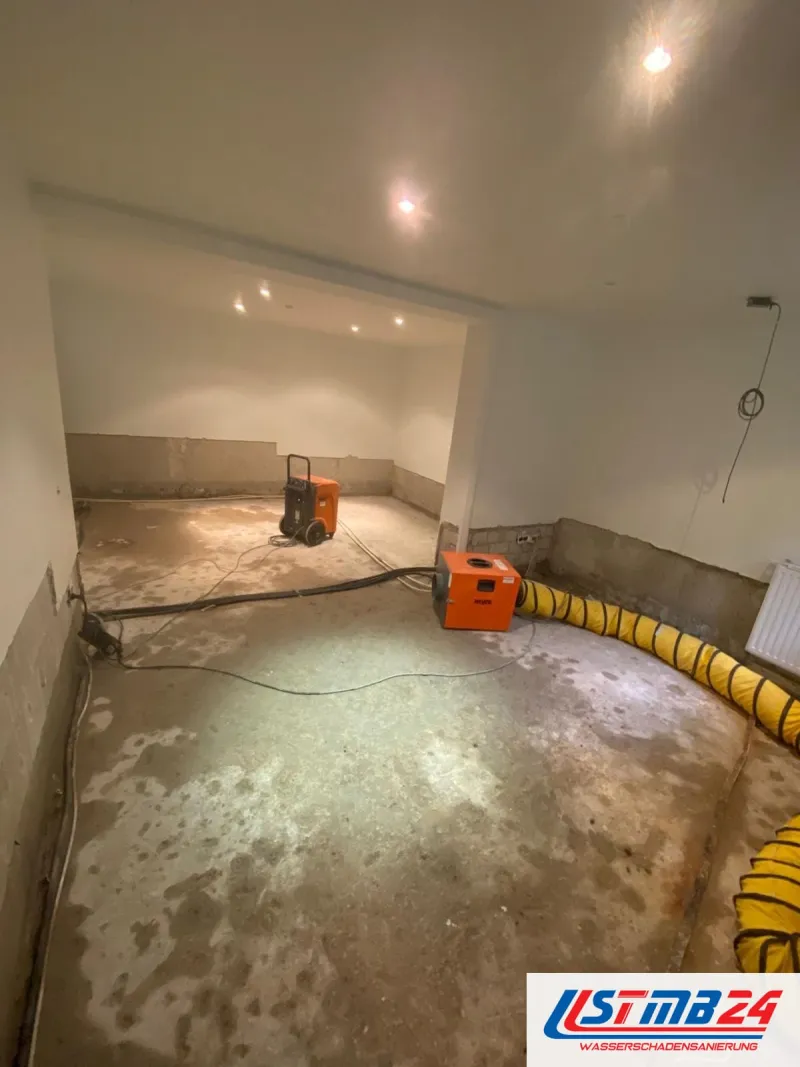 Bautrocknung in Grömitz: großer Souterrainraum, heller Estrich mit dunklen Wasserrändern, zwei Geräte und ein gelber Luftschlauch