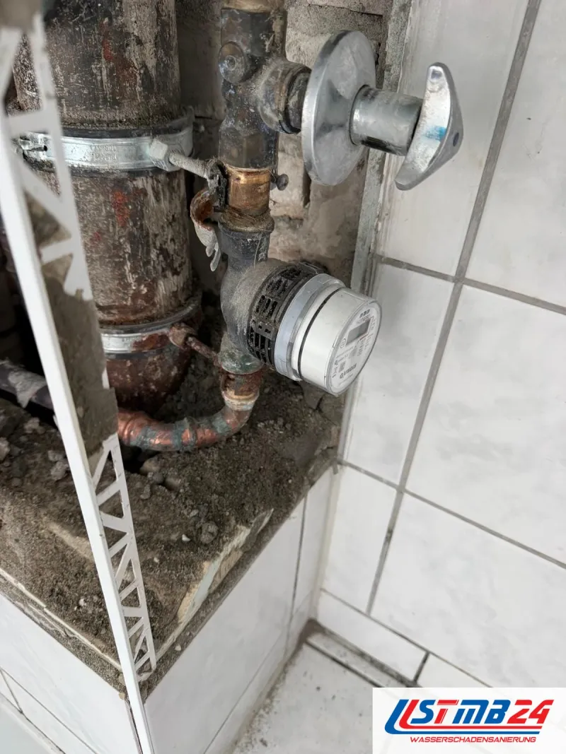 Sanitär und Heizung in Grömitz: Wasserzähler, Absperrventil und Gussrohr in der geöffneten Wand neben weißen Fliesen