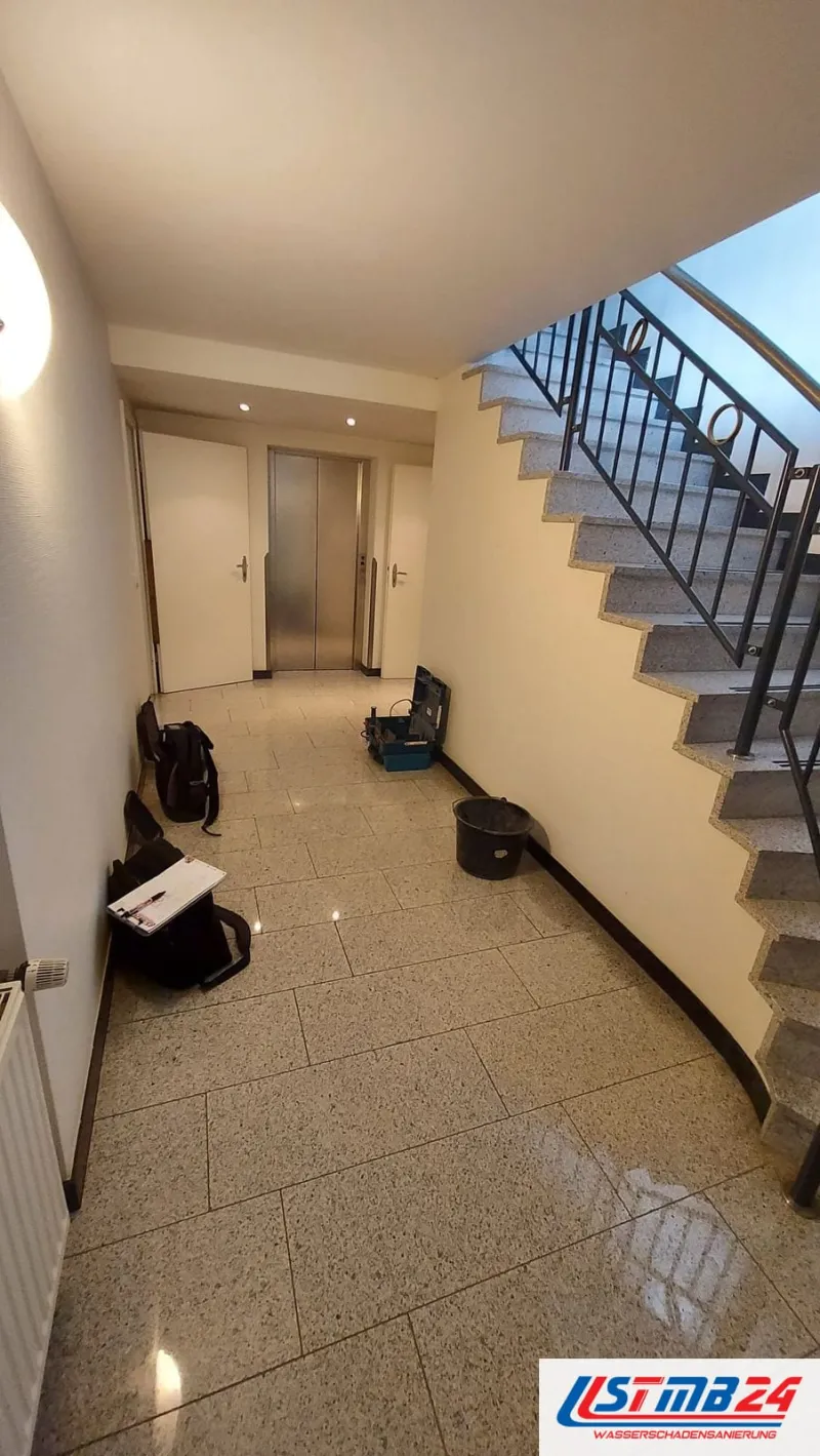 Vor der Trocknung: Treppenhaus mit nassem Steinboden, im Flur stehen Messkoffer, Taschen, ein Eimer und ein Klemmbrett