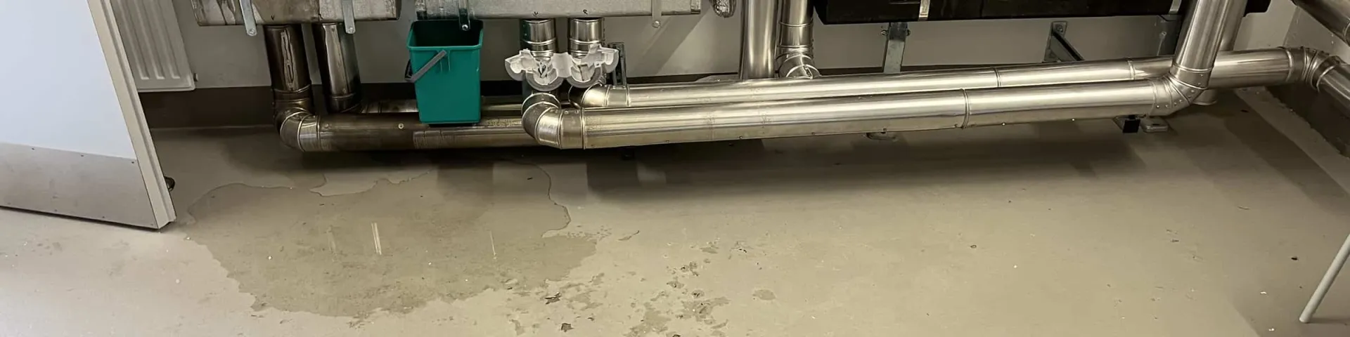 Trocknung: Wasserlache auf dem Boden eines Technikraums unter isolierten Rohrleitungen