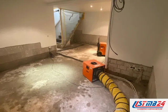 Trocknung im Keller: Trocknungsgerät mit gelbem Luftschlauch und Bautrockner auf dem freigelegten Estrich, Wandsockel bis aufs Mauerwerk geöffnet