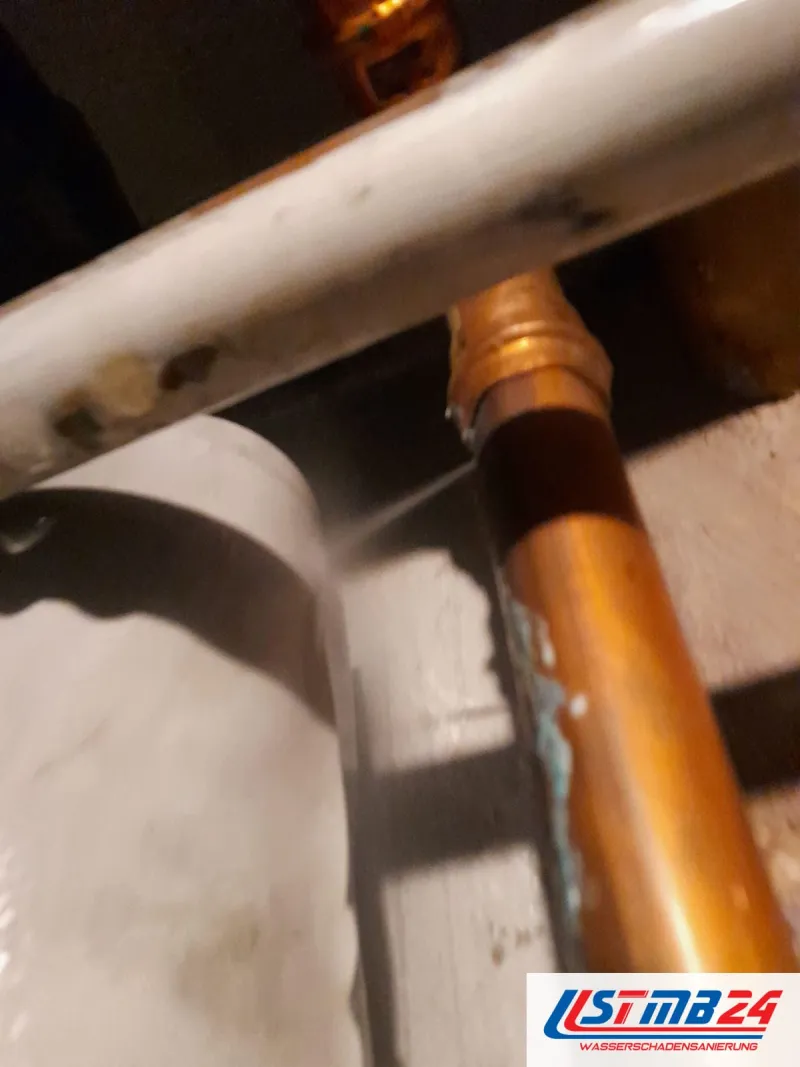 Ursachen von Wasserschäden: feiner Wasserstrahl aus einem Kupferrohr im Keller, am Rohr darunter Grünspan